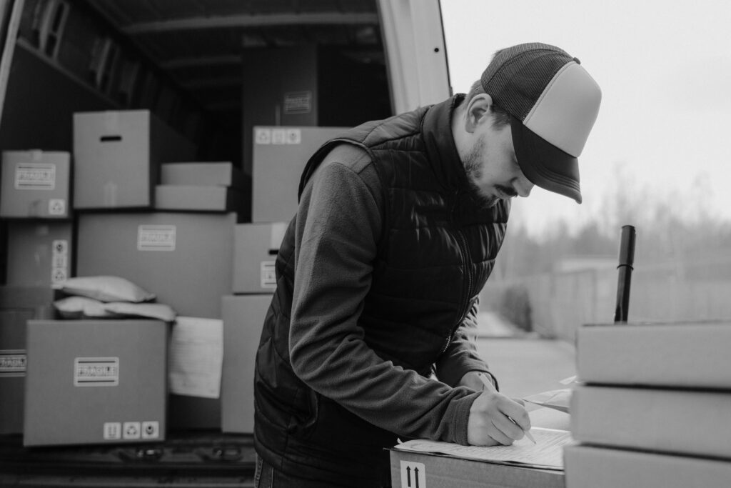 pexels photo 6169048 6169048 Black-and-white image of a courier documenting package details by a delivery van.